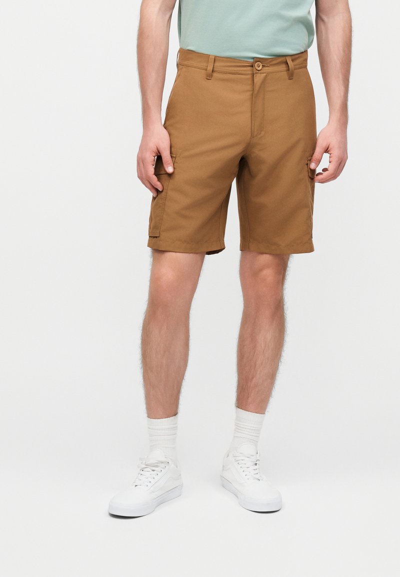 Άνδρας που φορεί καφέ βερμούδα με τσέπες έως το γόνατο, λευκές κάλτσες και λευκά αθλητικά παπούτσια, στέκεται μπροστά σε ένα απλό λευκό φόντο.