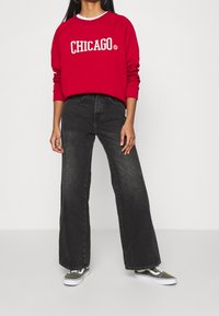 Felpa rossa con la scritta "CHICAGO", abbinata a jeans neri a vita alta e gamba larga, e scarpe da ginnastica verdi con suole bianche.
