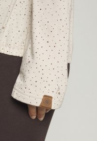 Light beige long-sleeve top with small brown polka dots, featuring a textured fabric and a branded leather tag on the cuff.