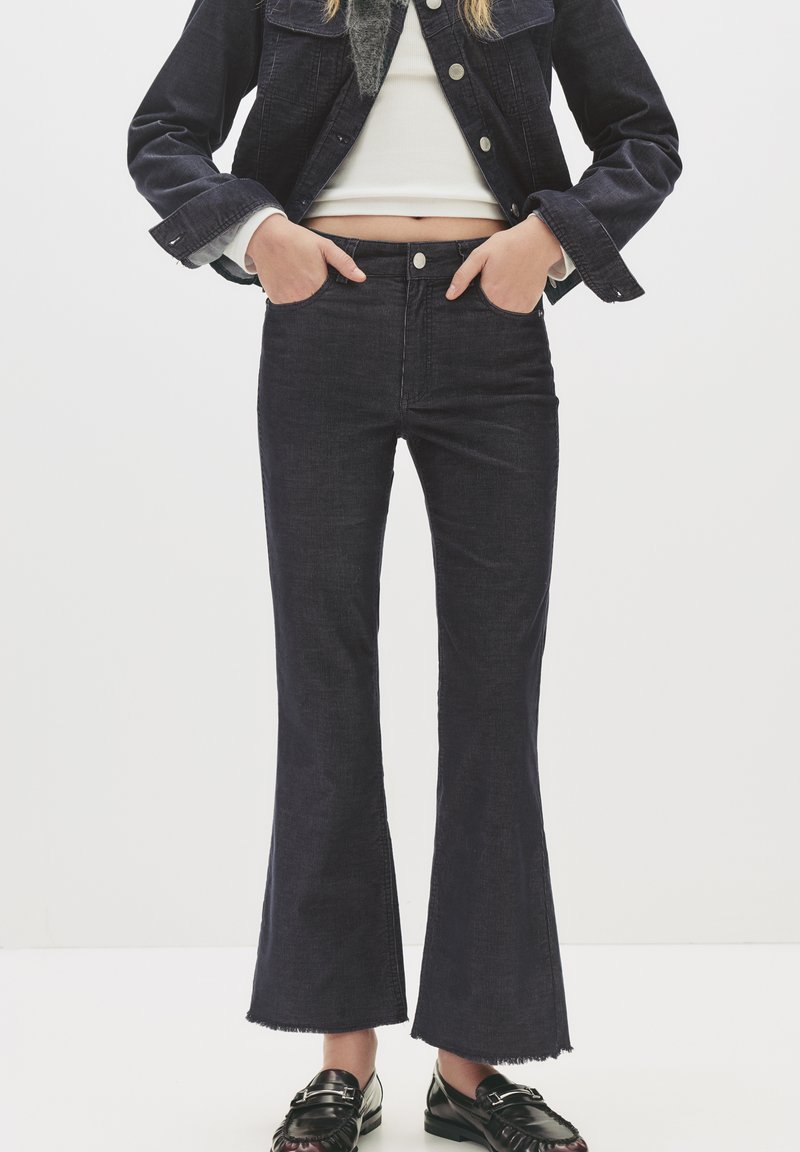 Ženska v črnih razširjenih kratkih hlačah, črni jakni, belem kratkem topu in črnih loaferjih, stoji z rokama v žepih.