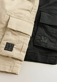 Twee paren cargoshorts, één in beige en één in zwart. Elk heeft meerdere zakken en een label met productdetails eraan bevestigd.