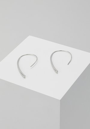 Boucles d'oreilles créoles en argent avec un design incurvé et minimaliste. Fabriquées en métal, présentant une texture lisse et une forme ouverte, exposées sur un socle blanc.