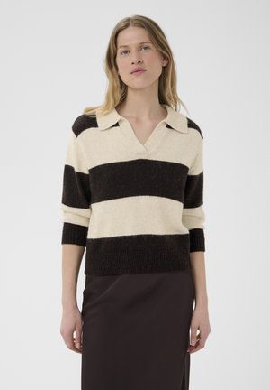Femme portant un pull à larges rayures noires et crème avec un col et une jupe foncée, debout devant un fond clair uni.