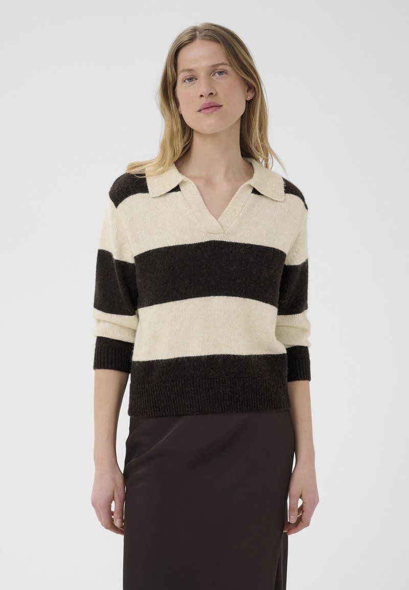 Femme portant un pull à larges rayures noires et crème avec un col et une jupe foncée, debout devant un fond clair uni.