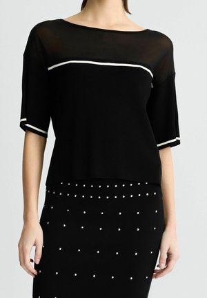 Kobieta ubrana w czarną bluzkę z krótkim rękawem z białym wykończeniem i prześwitującym dekoltem, w połączeniu z czarną spódnicą ozdobioną małymi białymi kropkami.