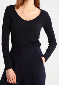 Top nero a maniche lunghe con scollatura a barca, realizzato in tessuto morbido e leggero; indossato con pantaloni scuri sartoriali.