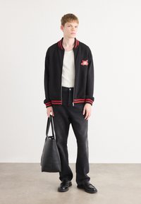 Black bomber jacket with red striped ribbing, white shirt, dark jeans, and black shoes. Model holds a black tote bag with a smooth texture.