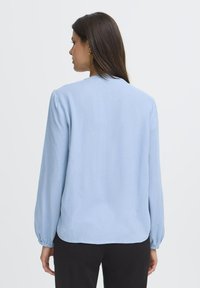 Femme aux longs cheveux noirs portant un blouse à manches longues bleu clair et un pantalon noir, de dos contre un fond blanc uni.