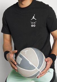 Svart t-shirt med kort ärm och vit Jordan-logga samt tryckt text. Håller en grå basketboll med vita detaljer och logga. Mintgröna shorts.