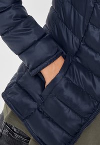 Giacca a vento blu navy con texture trapuntata, dotata di tasche laterali e una finitura liscia e lucida. Si sovrappone a una maglia verde, mostrando un look informale.