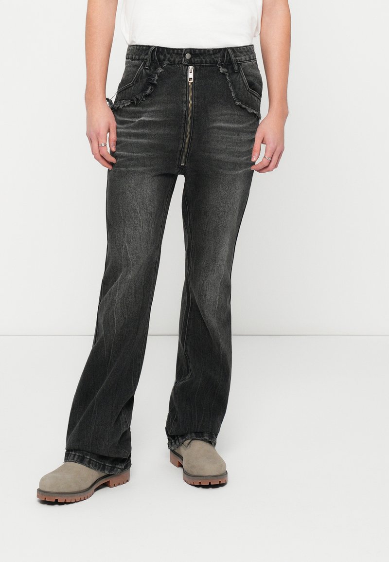 Calças de ganga pretas de cintura alta e boca de sino com detalhes desgastados, fecho à frente com zipper e dois bolsos laterais. Combinadas com uns sapatos cinzentos.