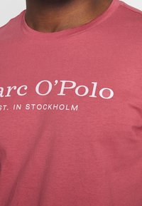 Bordowa koszulka z miękkiego materiału, z białym napisem "Marc O'Polo EST. W Sztokholmie" na piersi w nowoczesnej czcionce.