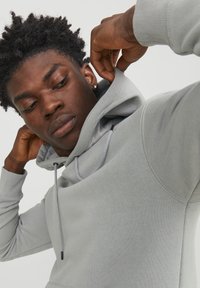 Light grey hoodie with a drawstring, soft texture, and kangaroo pocket; model adjusting the hood, showcasing a relaxed fit design.