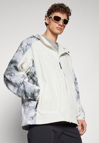 Lightweight, cream-colored jacket featuring a gray marble-patterned design on the sleeves, with a zip closure and hood. Accented with pockets.