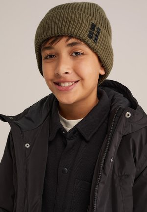 Un bonnet en ribbed vert olive avec un logo, porté avec un manteau à capuche noir et une chemise texturée noire en dessous, sur un fond neutre.