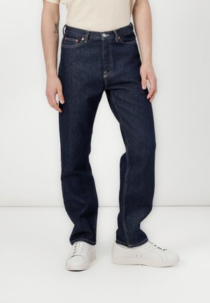 Mørkeblå denimjeans med lige ben, kontrastsyning og metalknaplukning i taljen. Bæres med hvide sneakers.