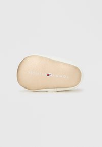 Μαλακή σόλα βρεφικού παπουτσιού σε μπεζ με λευκή περίμετρο, με το λογότυπο Tommy Hilfiger και το όνομα της μάρκας σε λευκό, με λεπτομέρεια σε κόκκινο-μπλε στο κέντρο.