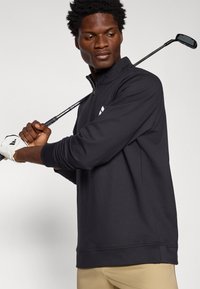 Black pullover sweater with a high collar and long sleeves. Made of smooth fabric. Features a subtle logo on the left chest. Worn with a golf club.
