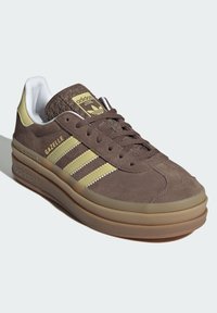 Brune semskede sneakers med cremefargede aksenter og tre lysegrønne striper. Har en teksturert brun tunge og en tykk gummisåle.
