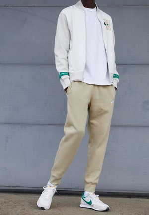 Personne portant un pantalon de survêtement beige Nike, des baskets blanches Nike avec le logo Swoosh vert, un t-shirt blanc et une veste blanche avec des bandes vertes et le logo N.
