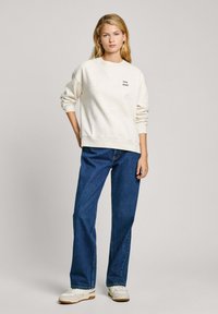 Kreminės spalvos džemperis su dėmėta tekstūra, su juodu „PEPE JEANS“ logotipu, dėvimas su laisvomis mėlynomis džinsinėmis kelnėmis ir baltomis sportinėmis bateliais.
