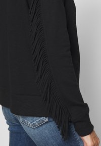 Černá mikina s texturovanými třásněmi podél jedné strany, v kombinaci s modrými denimovými džíny, předvádí pohodlný a vrstvený vzhled.