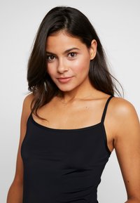 Black fitted camisole top with thin straps, smooth texture, and a rounded neckline. Worn by a person with long dark hair.