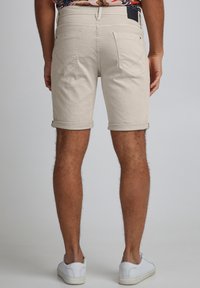 Pantaloncini di cotone beige con risvolti, due tasche posteriori e una piccola etichetta nera del marchio in vita, abbinati a scarpe da ginnastica bianche.
