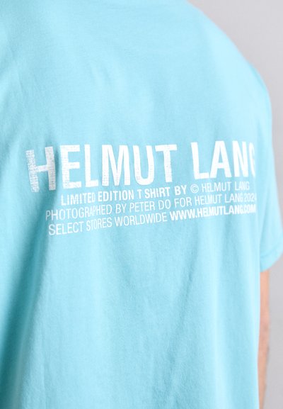 Svijetloplava pamučna majica s bijelim natpisom na leđima, s nazivom branda i detaljima o limitiranom izdanju i fotografu.