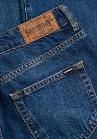 Close-up af blå denimjeans baglomme med orange syninger og brun læder Superdry-mærke over lommen.