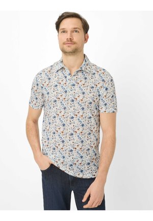 Uomo che indossa una camicia a maniche corte bianca con motivi floreali blu e marroni, in piedi con una mano in tasca.