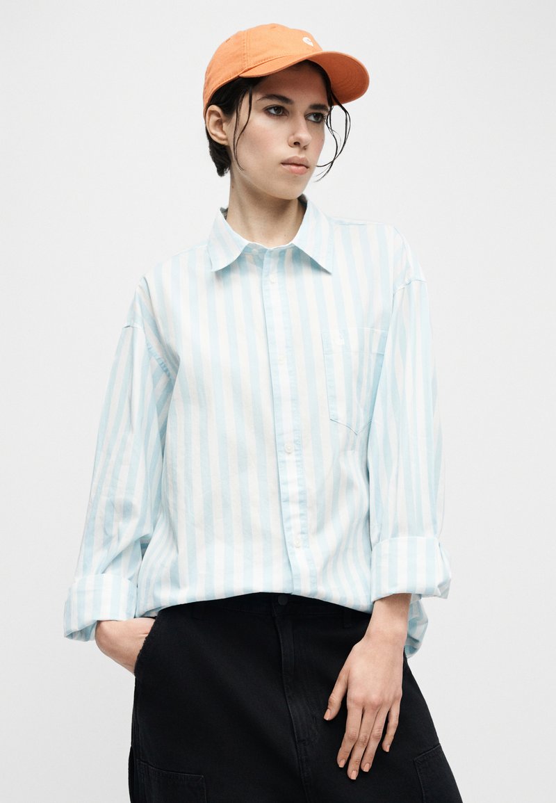 Giovane con indosso un cappellino arancione, camicia a righe azzurre e bianche con bottoni e pantaloni neri, in piedi con una mano in tasca.