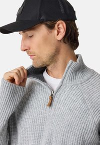 Man wearing black cap and light gray ribbed zip sweater, pulling collar up to reveal dark gray inner lining.