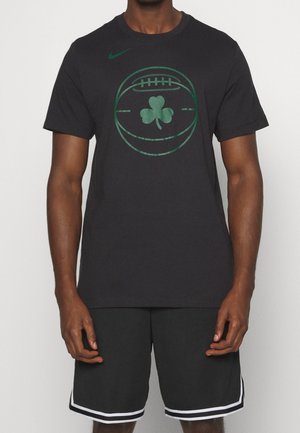 Man draagt zwarte shorts en een zwart t-shirt met een groene basketbal- en klavergrafiek in het midden en een klein groen Nike-logo.