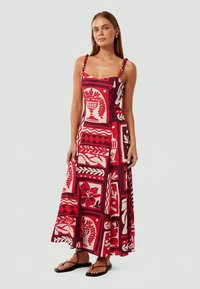 Mulher a usar um vestido sem mangas, até ao tornozelo, com padrões florais e geométricos em vermelho, rosa e bordô, com alças finas de contas.
