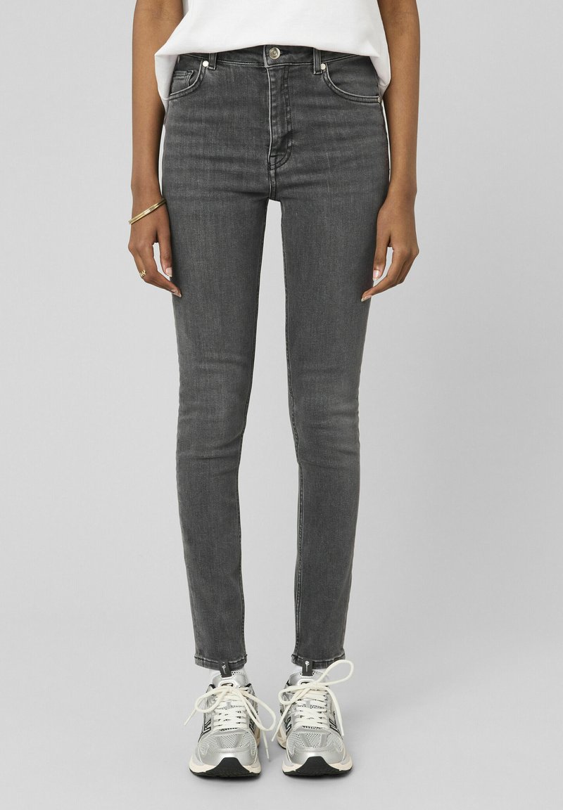 Personne portant un jean noir délavé slim, des baskets blanches avec des accents noirs et argentés, une chemise blanche rentrée, et des bijoux dorés à la main gauche.