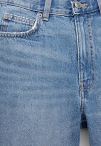 Gros plan d'un jean en denim bleu clair montrant les poches avant, les passants de ceinture, la fermeture par bouton et les détails de couture orange.