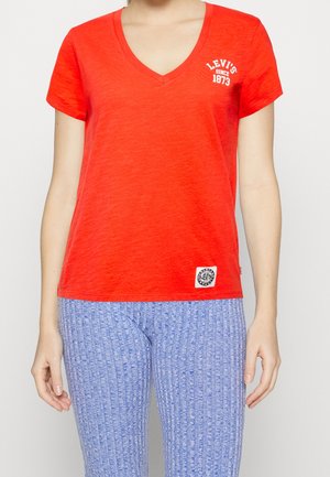 Nainen, jolla on päällään punainen V-kaula-aukollinen Levi's-T-paita valkoisella logotekstillä ja vaaleansiniset teksturoidut housut, seisoo yksiväristä taustaa vasten.