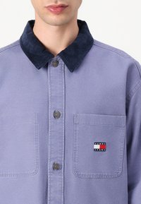 Faded lavender shirt with a navy corduroy collar, front metal buttons, and a left chest pocket featuring a small red, white, and blue logo.