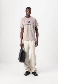 Beige cotton t-shirt featuring a black "BELSTAFF" logo, paired with light cream trousers and holding a black leather bag.