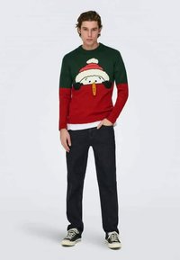Red and green sweater with snowman design, white and red hat, black buttons, worn with black jeans and black sneakers. Smooth knit fabric.