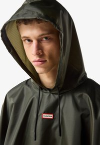Νέος άνδρας με γαλανά μάτια που φοράει σκούρο πράσινο αδιάβροχο μπουφάν με κουκούλα και λογότυπο «Hunter» στο στήθος.