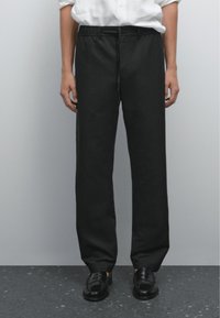 Persona con pantaloni neri e scarpe nere, in piedi contro uno sfondo neutro, mani ai lati, con una camicia bianca parzialmente infilata dentro.