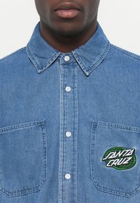 Denim košulja svijetloplave boje s raširenim ovratnikom, prednjim zatvaranjem na gumbe, dva džepa na prsima i vezom u crnoj i zelenoj boji.