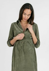 Olijfgroene corduroy jurk met een kraag, lange mouwen en een elastische tailleband. De stof heeft een verticale ribbelstructuur.