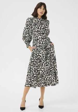 Robe midi à motifs léopard avec une taille cintrée, des manches longues et des poches. Le tissu est lisse avec des couleurs noir et beige. Des escarpins noirs complètent le look.