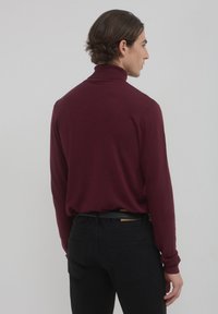Maglione a collo alto color bordeaux con maniche lunghe, texture morbida e design aderente. Indossato con pantaloni neri, che mostrano sottili dettagli delle cuciture posteriori.