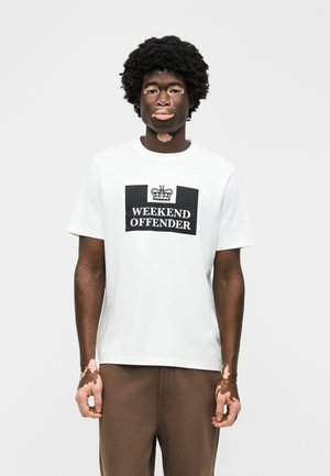 Νεαρός άνδρας με λεύκη που φοράει λευκό μπλουζάκι "Weekend Offender" και καφέ παντελόνι, στέκεται μπροστά σε απλό ανοιχτό γκρι φόντο.
