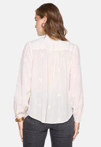 Blusa bianca a maniche lunghe in tessuto testurizzato, con retro plissettato e sottile motivo floreale. Orlo arrotondato che si ferma appena sopra i fianchi.