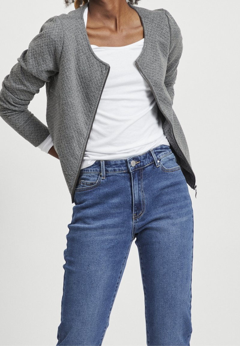 Personne portant une chemise blanche, une veste zippée texturée grise et un jean en denim bleu, montrée du cou aux hanches sur un fond uni.